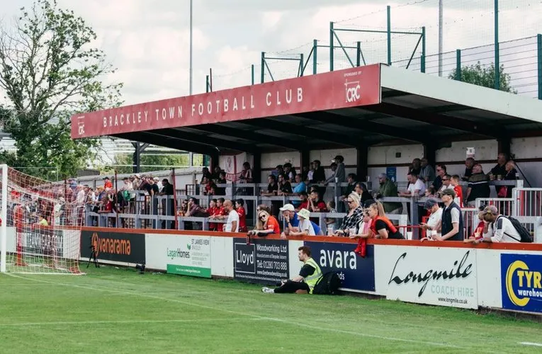Khung cảnh sân St James Park Brackley trong ngày thi đấu, với không khí sôi động của người hâm mộ bóng đá địa phương.